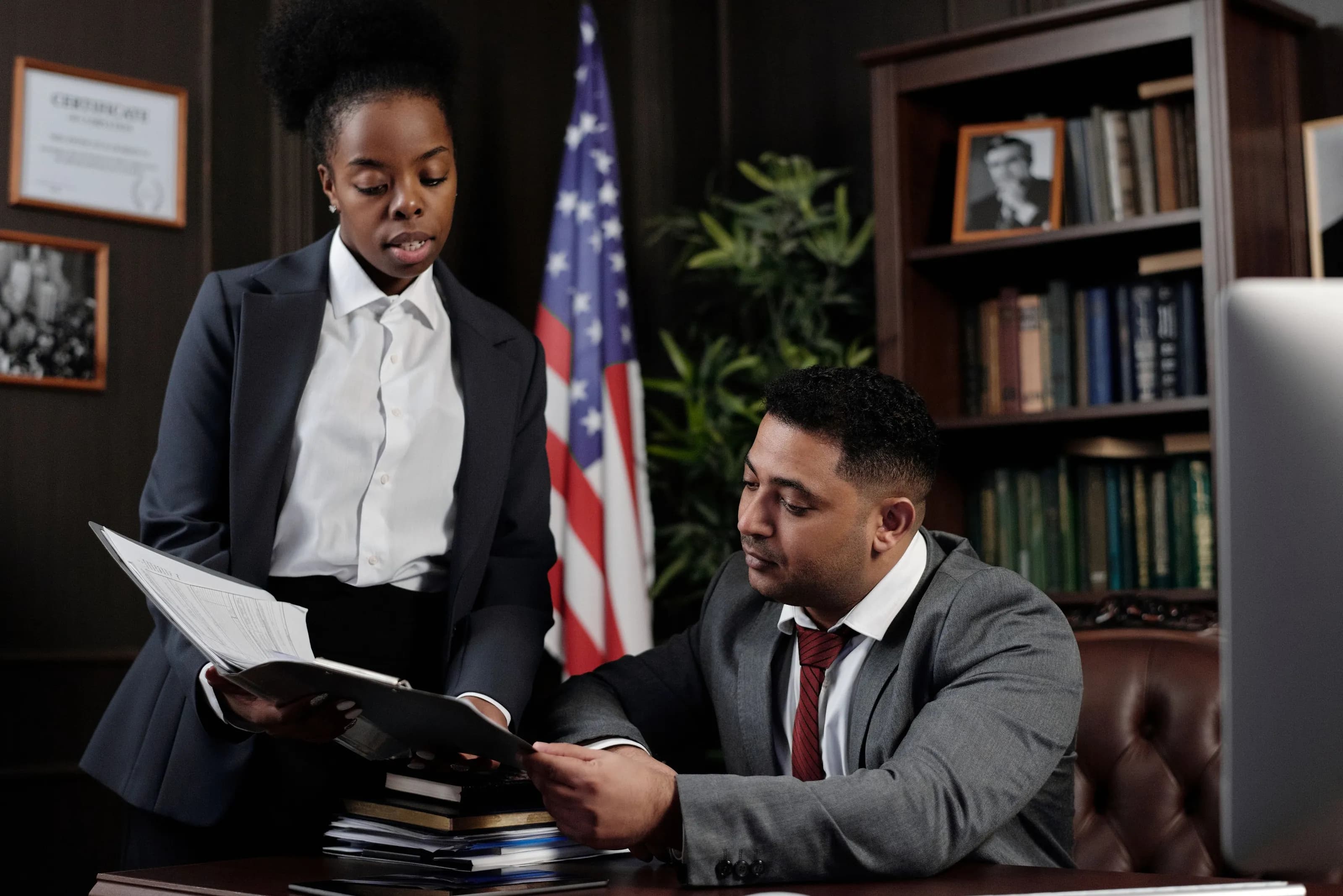 A lawyer stands at a colleague's desk and walks him through a document in a wood-paneled office.