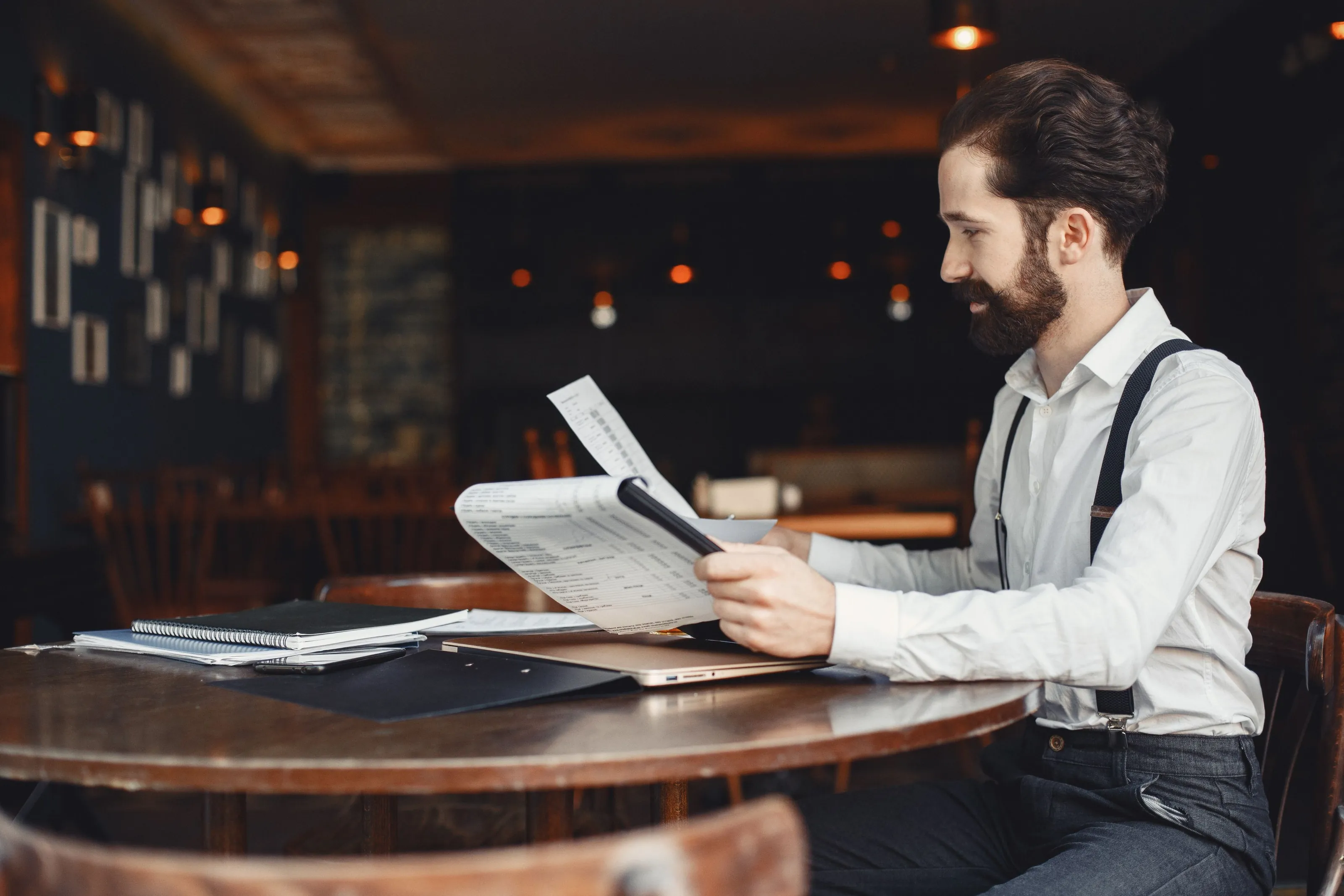 A man in shirtsleeves reads a printout at a table in an empty, dimly lit restaurant.