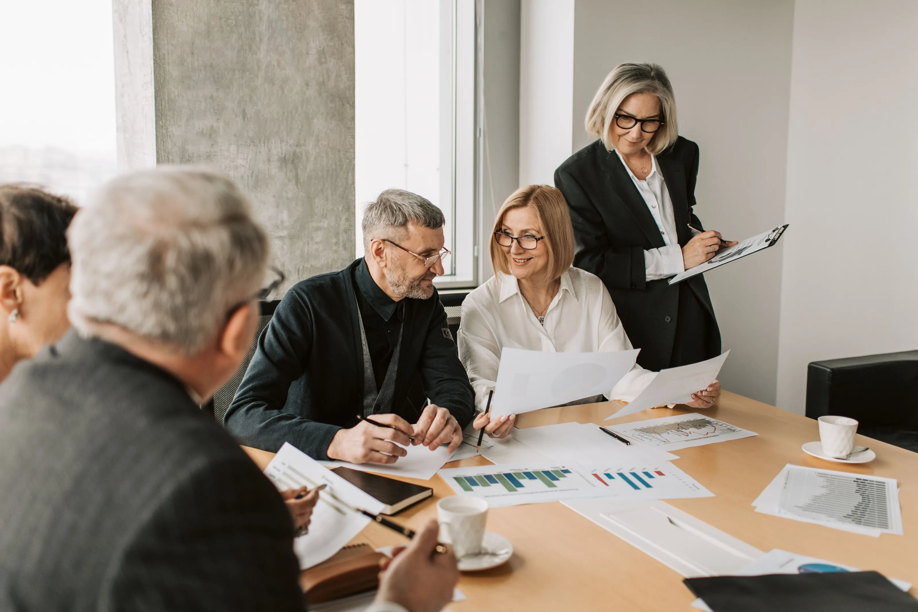 Four colleagues around a conference table covered in printed schedules. One stands with a clipboard and points at a page.